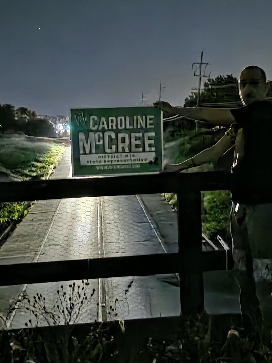 Rob Gryder holds a McCree sign at arm's length over the railing of the Route 47 rail overpass in Yorkville at night; headlights from the Boombah Lane intersection light the sign from behind.