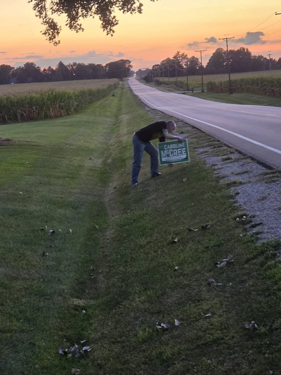 A person bends over a McCree sign beside a rural road at sunset.